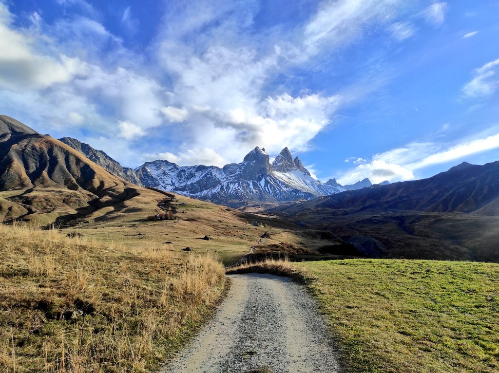 Chemin de gravier au milieu des prairies d'automne, filant droit vers les Aiguilles d'Arves saupoudrées de neige sous un ciel nuageux