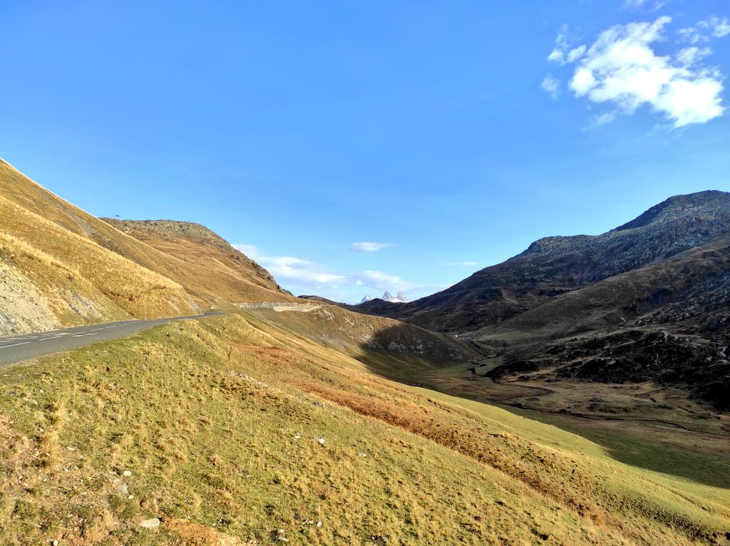 Route du col de la Croix de Fer entre des pentes d'herbe dorée, avec au loin, au creux de la vallée, les trois pointes enneigées des Aiguilles d'Arves sous un ciel bleu