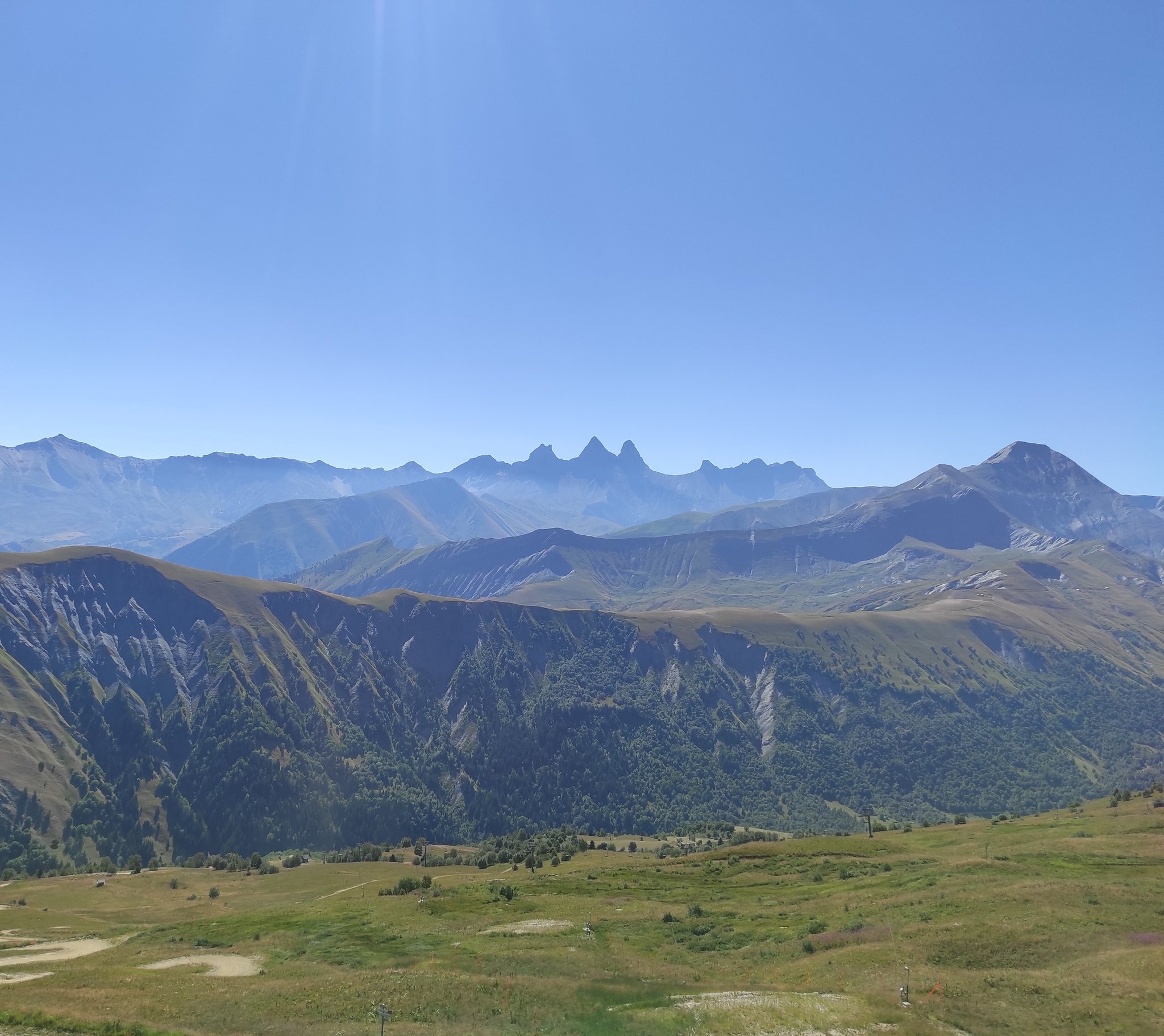 Panorama d'été sur les alpages et les crêtes boisées, avec à l'horizon la silhouette des trois Aiguilles d'Arves dont les pointes rappellent des oreilles de chat