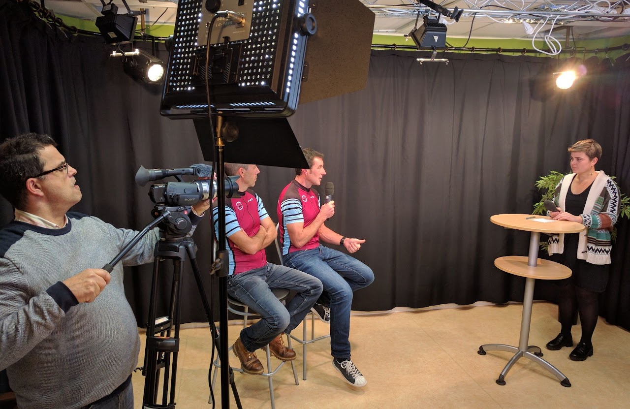 2016, Anne, bénévole, interviewe deux hommes en tenue de cyclisme assis sur des tabourets dans le studio, sous les projecteurs.