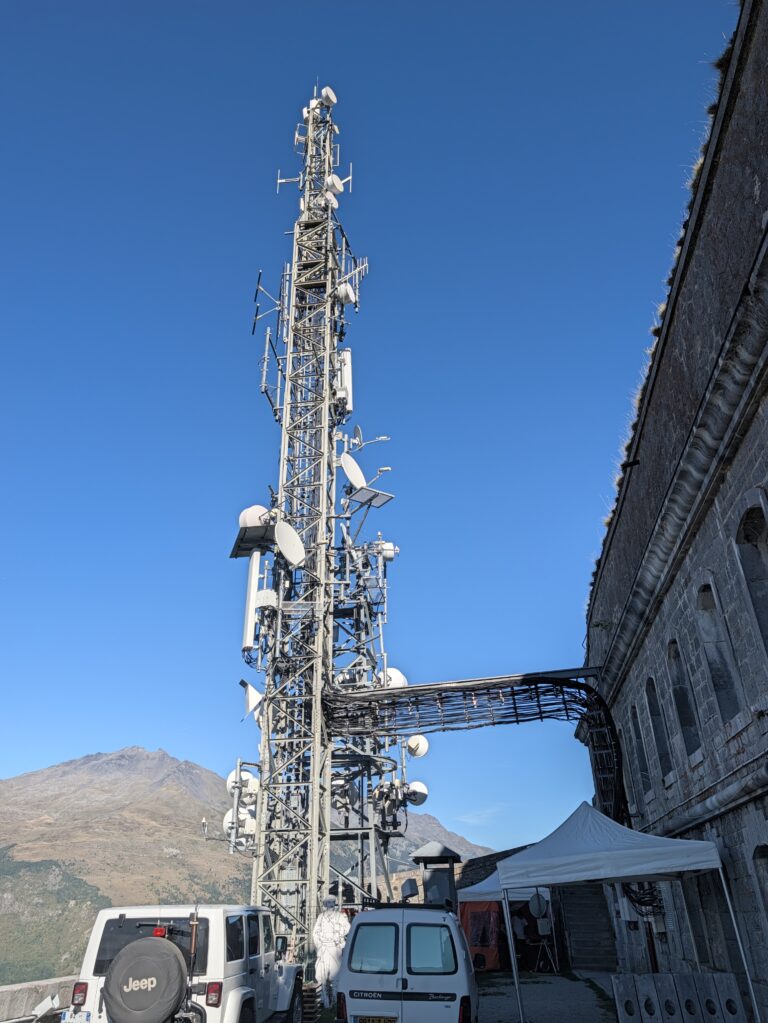 Pylône de télécommunications hérissé d'antennes et de paraboles, accolé au bâtiment en pierre du Fort du Télégraphe à Valloire, avec une montagne en arrière-plan sous un ciel bleu et des véhicules garés au pied de la tour