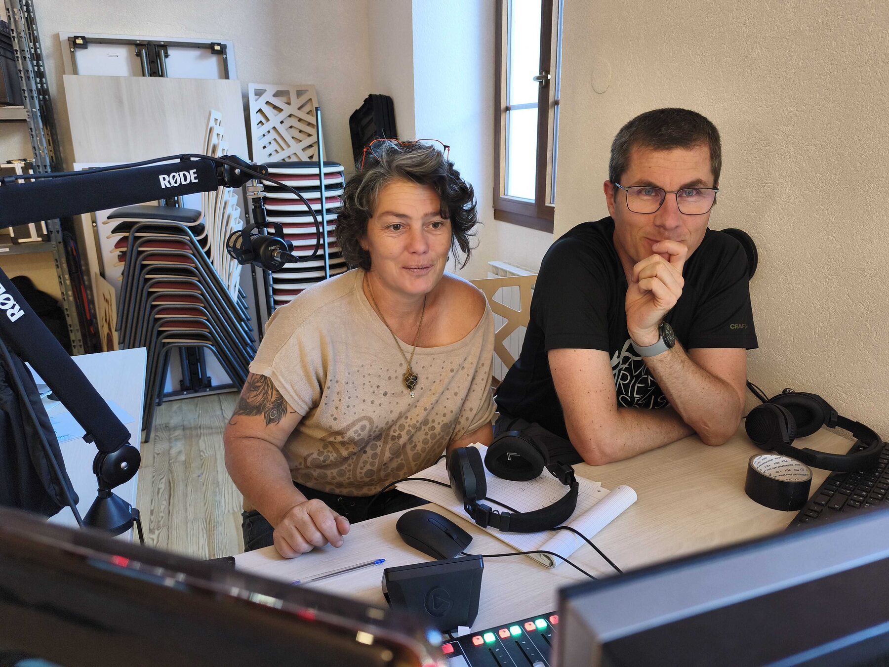 Deux bénévoles du studio MAUZ à la régie, concentrés sur les écrans, avec un micro Rode, des casques et la table de mixage sur le bureau