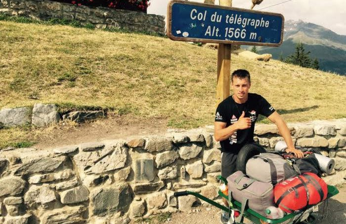 2018, un homme pose avec une brouette chargée de sacs, devant le panneau du col du Télégraphe (alt. 1566 m), lors du tour de la Maurienne en brouette.