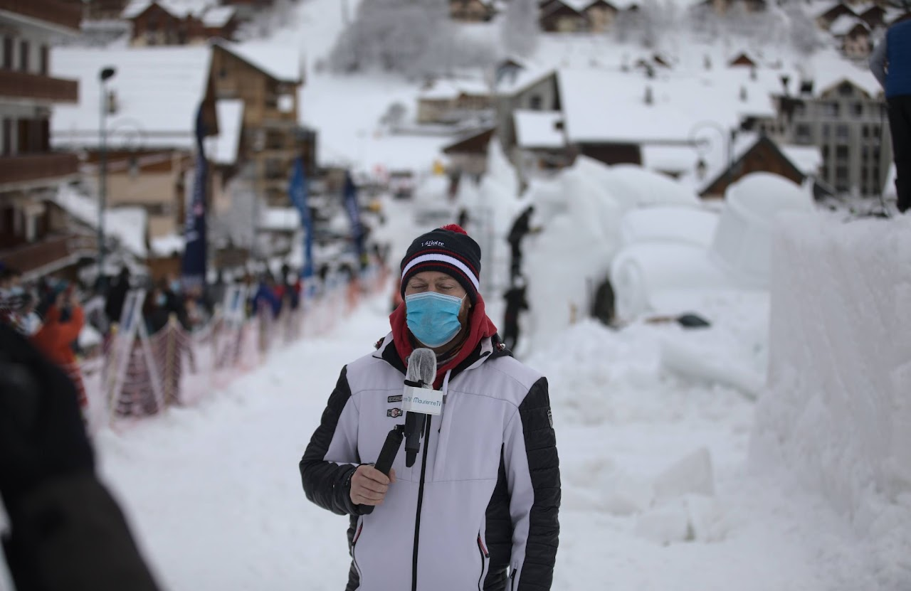 Hiver 2021, Cédric en reportage à Valloire, masqué, micro Maurienne TV à la main, sculptures de neige et station enneigée en fond.