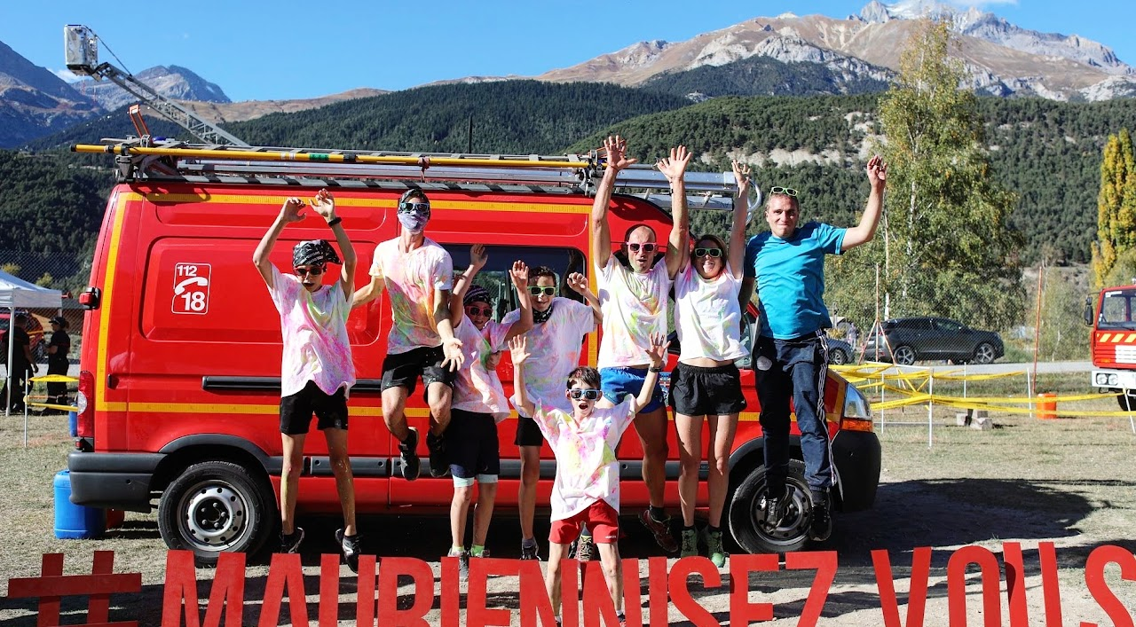 2018, un groupe de participants couverts de poudre colorée saute devant un camion de pompiers, montagnes en arrière-plan, lors d'un color run.