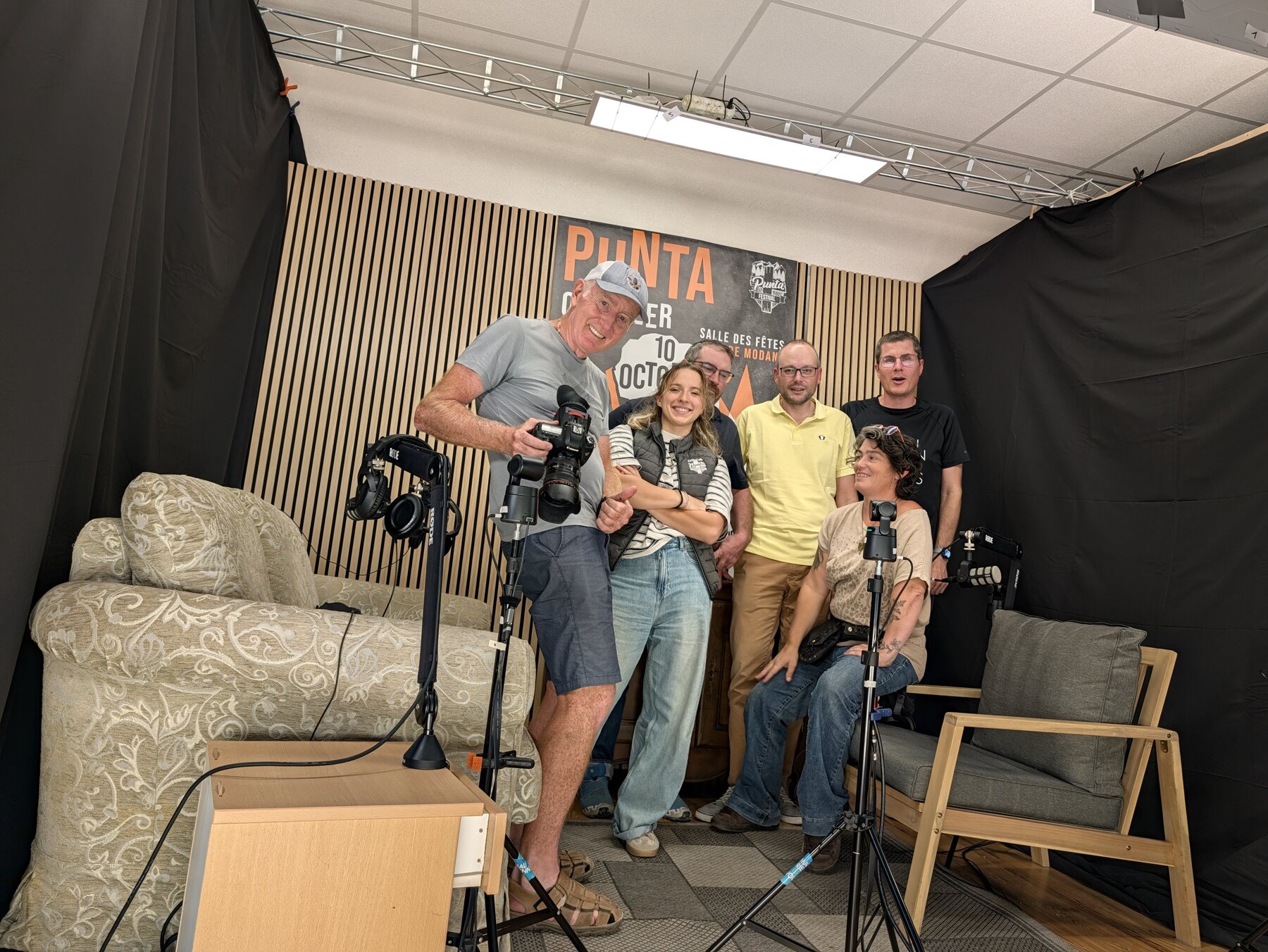 Les invités de Punt'assoc et les bénévoles du studio MAUZ posent dans le studio, devant l'affiche du Punta Octobeer Festival, avec caméras et micros autour du canapé