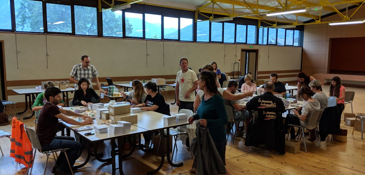 2018, des bénévoles assis à plusieurs tables dans une salle des fêtes préparent la distribution des 25 000 autocollants #MauriennisezVous.