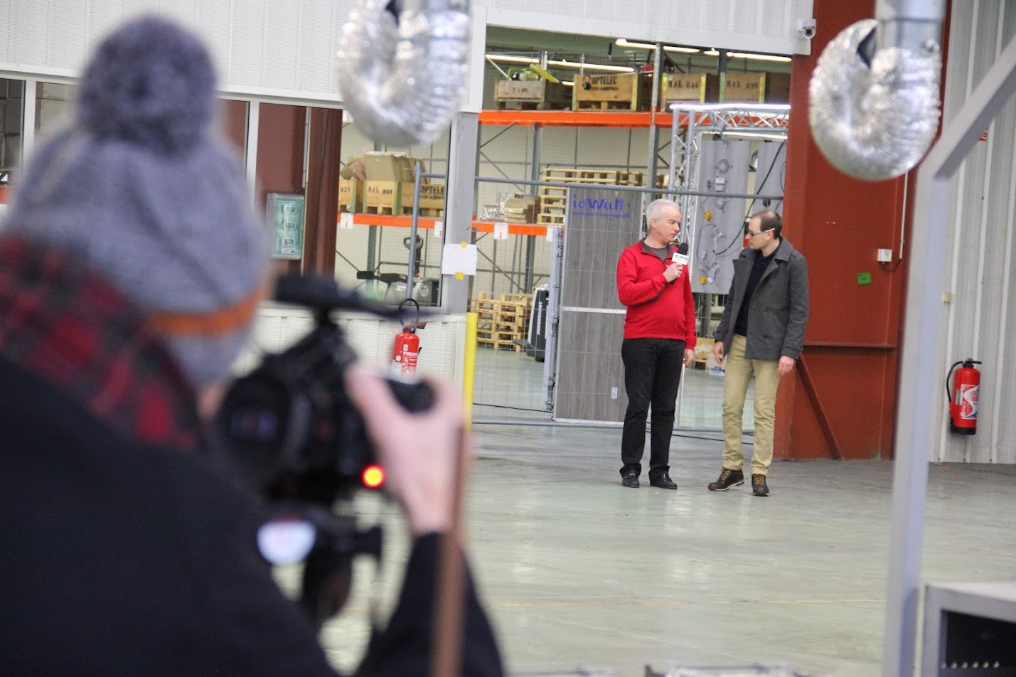 2016, Florian interviewe deux hommes dans un entrepôt industriel de l'entreprise IdWatt, à Saint-Rémy-de-Maurienne, un caméraman au premier plan.