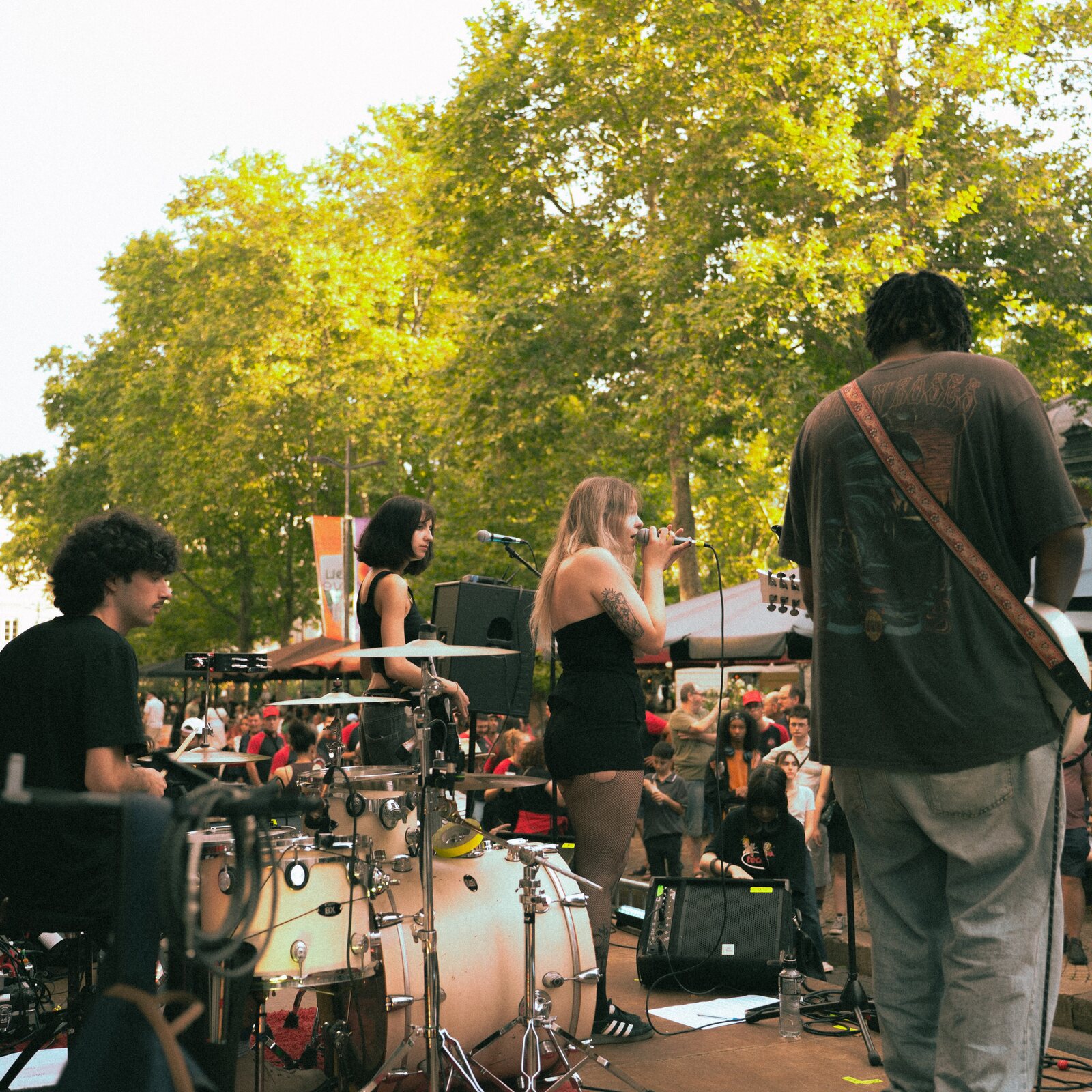 Le groupe stéphanois 404 sur une scène en plein air : le batteur, une musicienne, la chanteuse au micro et un guitariste de dos, face au public rassemblé sous les arbres
