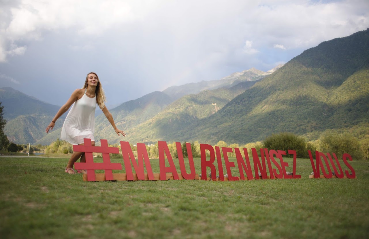 2018, une femme en robe blanche danse près des lettres géantes en bois #MAURIENNISEZ VOUS posées dans un pré, arc-en-ciel et montagnes en fond.