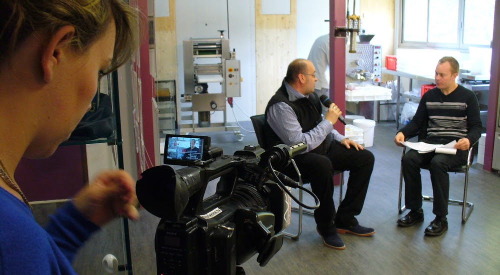 2014, Cédric interviewe un homme assis dans un atelier de fabrication artisanale, caméra au premier plan.