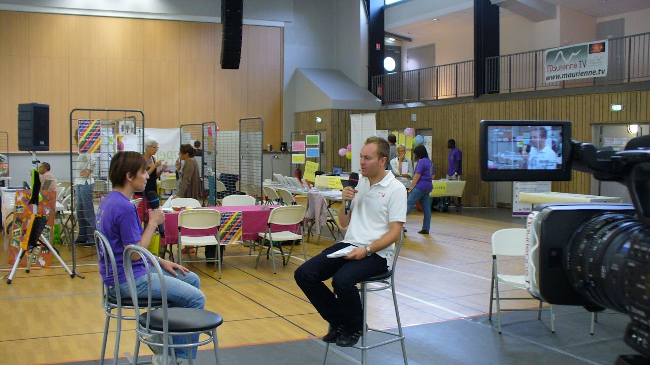 2013, interview de Cédric assis sur un tabouret dans une salle des fêtes lors d'un événement associatif, caméra et écran de retour au premier plan, banderole Maurienne TV en arrière-plan.