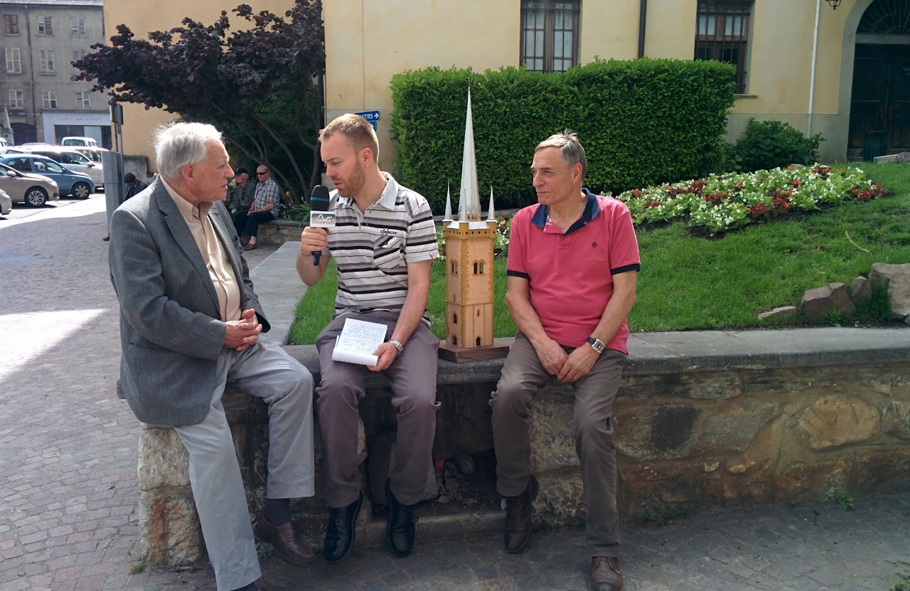 2015, Cédric interviewe deux hommes assis sur un muret en extérieur, une maquette de tour à côté d'eux.