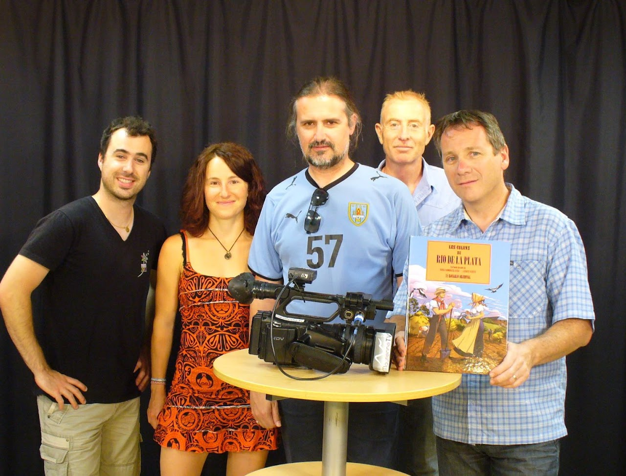 2014, l'équipe pose avec Mélina autour d'une caméra et de la bande dessinée « Les Colons de Rio de la Plata », fond noir de studio.