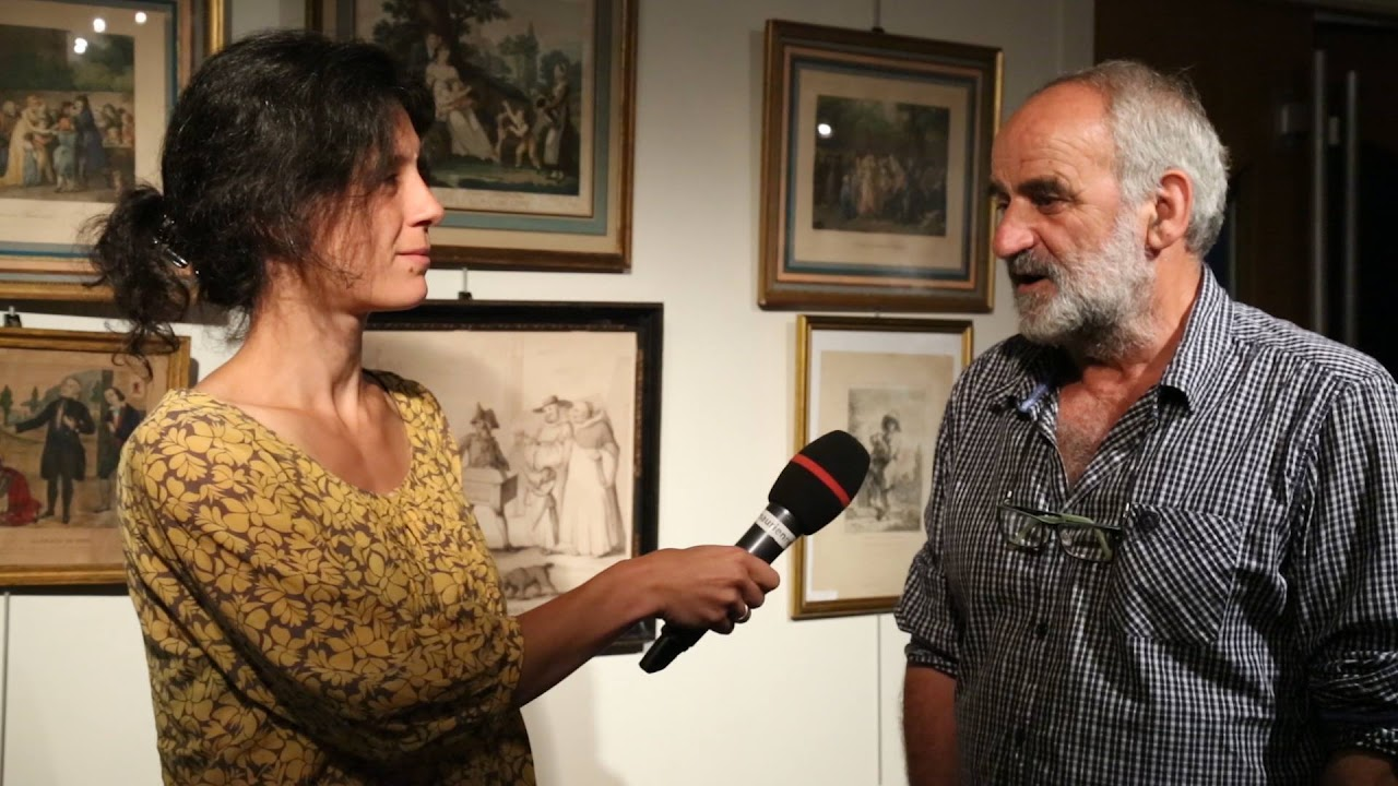 2016, Laetitia interviewe un homme au micro dans une salle décorée de gravures anciennes encadrées.