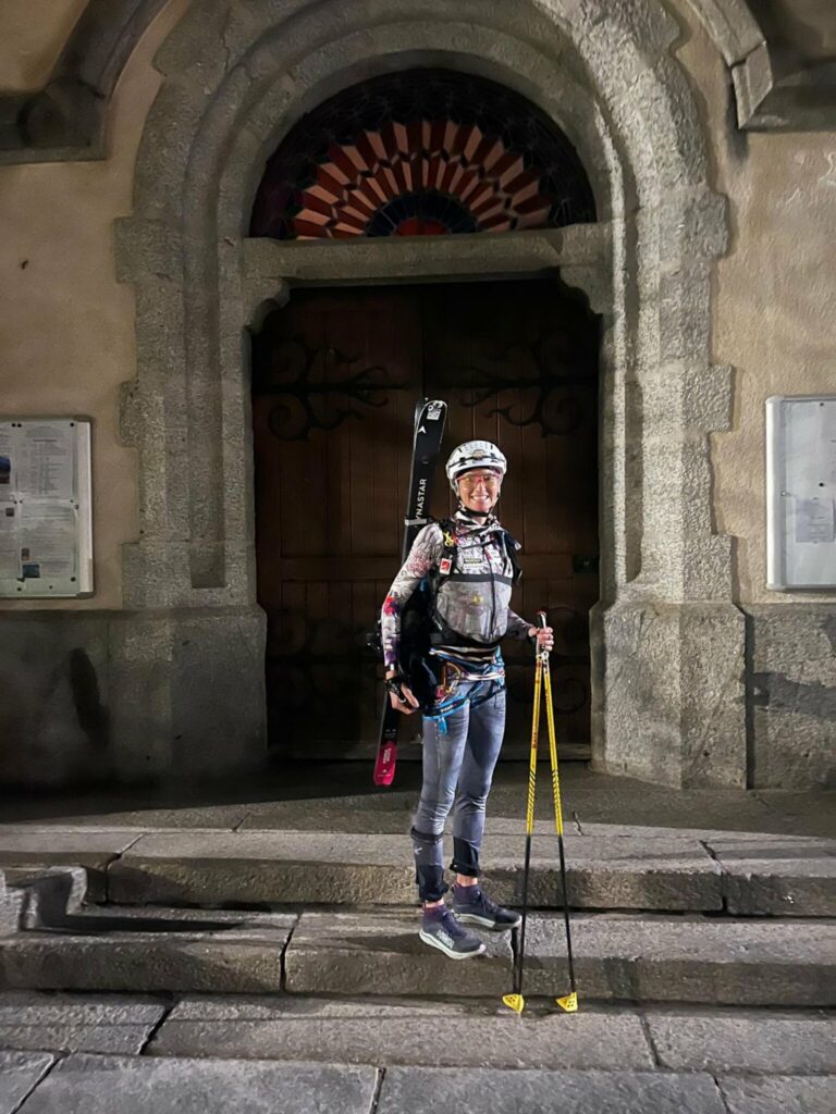 Lorna Bonnel debout sur les marches de l'église de Chamonix, skis sur le sac et bâtons jaunes à la main, avant le départ
