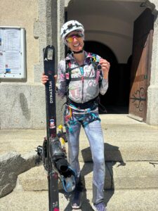 Lorna Bonnel, casque blanc et lunettes de soleil, sourit devant l'église de Chamonix avec un ski Dynastar à la main et sa montre de sport levée
