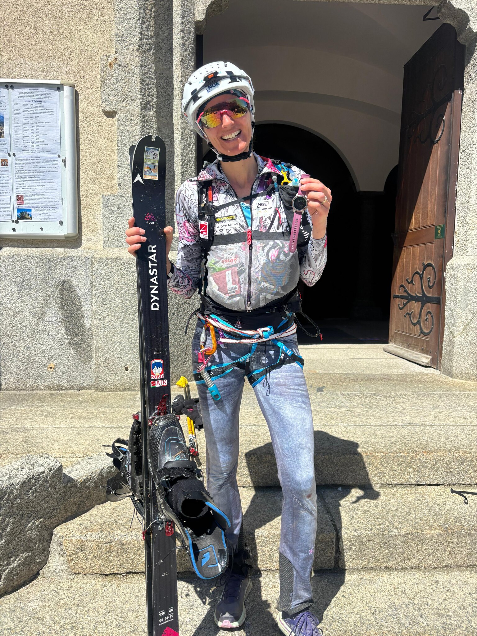 Lorna Bonnel, casque blanc et lunettes de soleil, sourit devant l'église de Chamonix avec un ski Dynastar à la main et sa montre de sport levée