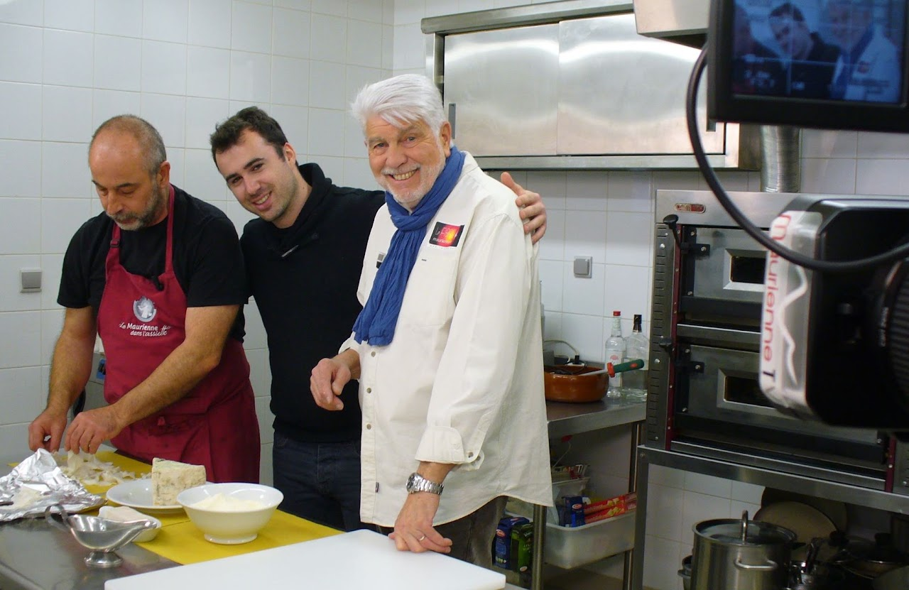 2015, tournage de « Maurienne à table » avec Jean-Claude : un cuisinier prépare des fromages en cuisine, aux côtés d'un autre membre de l'équipe, caméra Maurienne TV au premier plan.