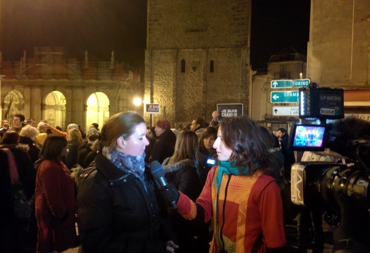 2015, Mélina interviewe une passante de nuit lors d'un rassemblement place centrale, pancartes « Je suis Charlie » visibles dans la foule.
