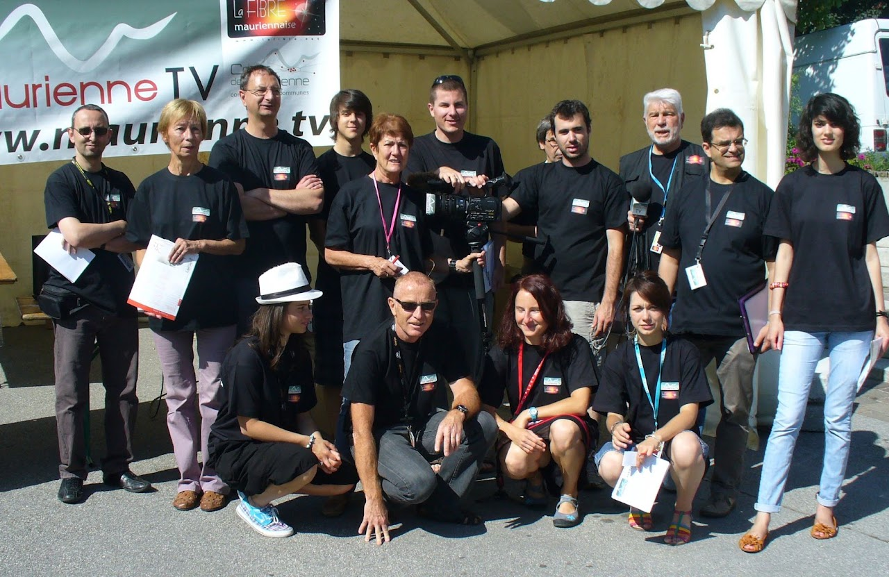 2012, l'équipe de Maurienne TV et La Fibre Mauriennaise au grand complet, en t-shirts noirs, posant sous la banderole de la chaîne lors d'une présence sur un marché ou une fête.