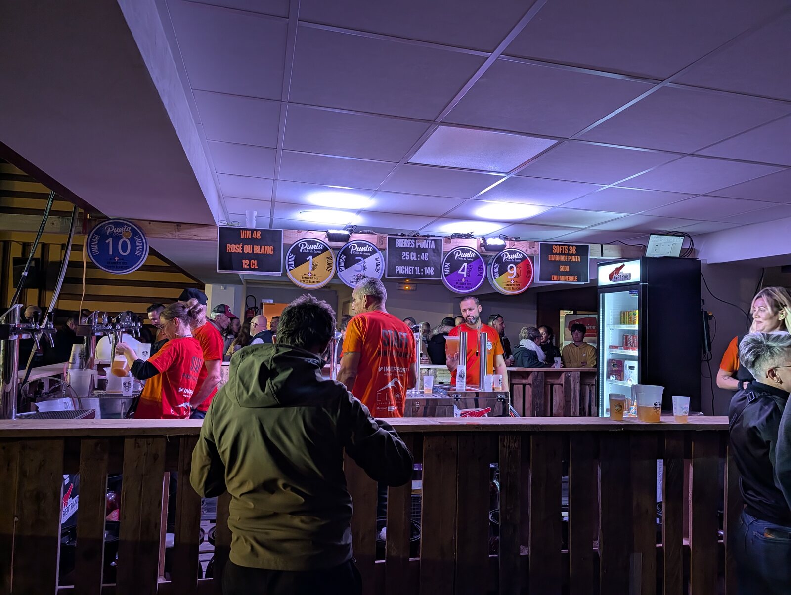 Le bar de la Punta Octobeer 2025 à Modane : des bénévoles en t-shirt rouge et orange tirent les bières de la Brasserie des Terres Blanches (blonde, blanche, NEIPA, mangue, limonade) sous les enseignes du festival