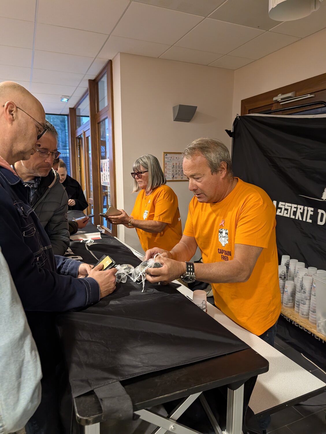 Deux bénévoles de Punt'assoc en t-shirt orange accueillent les festivaliers à l'entrée de la salle des fêtes de Modane et préparent des petits sachets, avec des éco-cups empilés à côté d'eux, Punta Octobeer 2025