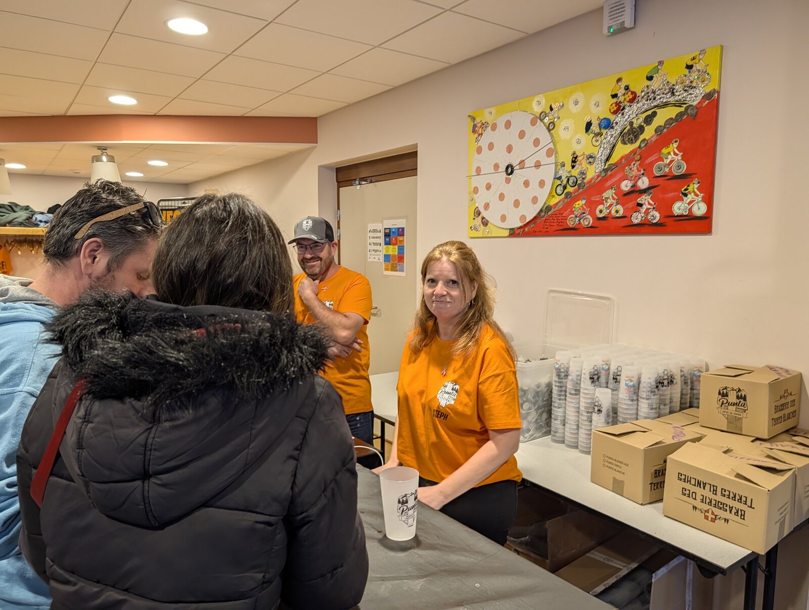 Deux bénévoles de Punt'assoc en t-shirt orange du festival sourient derrière leur table, avec des piles d'éco-cups et des cartons de la Brasserie des Terres Blanches, devant des festivaliers, Punta Octobeer 2025 à Modane
