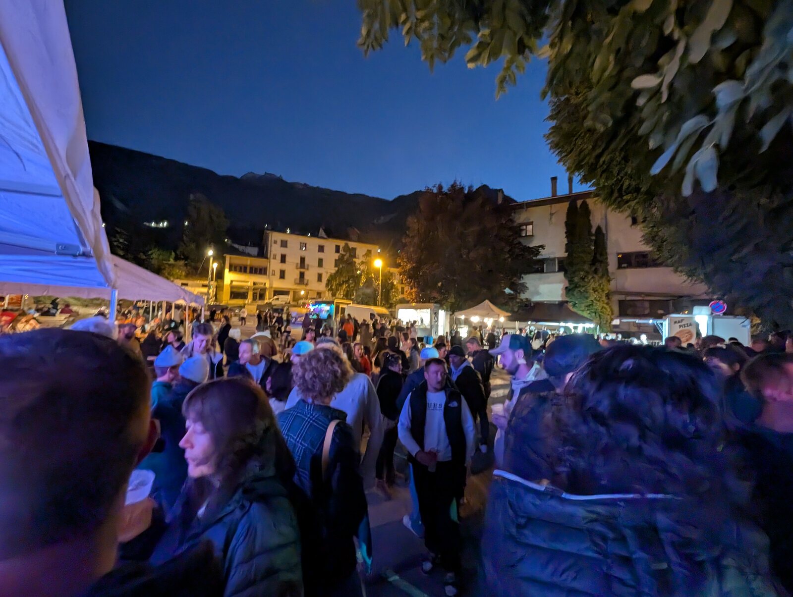 La foule des festivaliers de la Punta Octobeer 2025 sur la place de l'Europe à Modane à la tombée de la nuit, entre les chapiteaux et les food trucks illuminés, avec les montagnes en fond