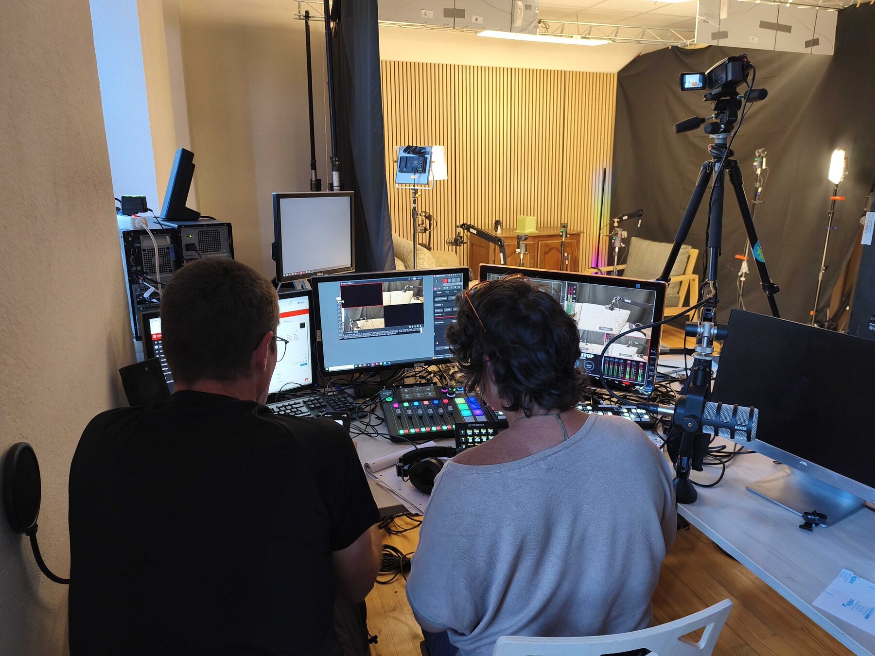 Deux bénévoles vus de dos devant les écrans et la table de mixage de la régie, le studio d'enregistrement éclairé en face d'eux