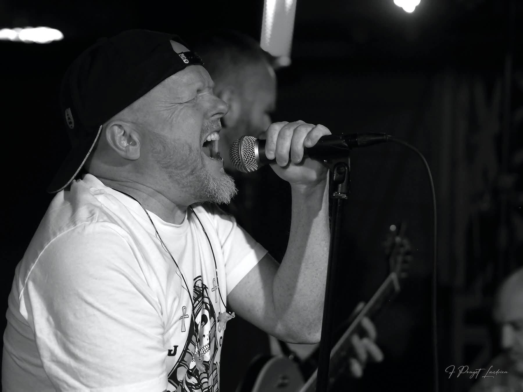Photo en noir et blanc du chanteur de Rockface en concert, casquette à l'envers, en plein chant devant un micro sur pied, avec un guitariste derrière lui