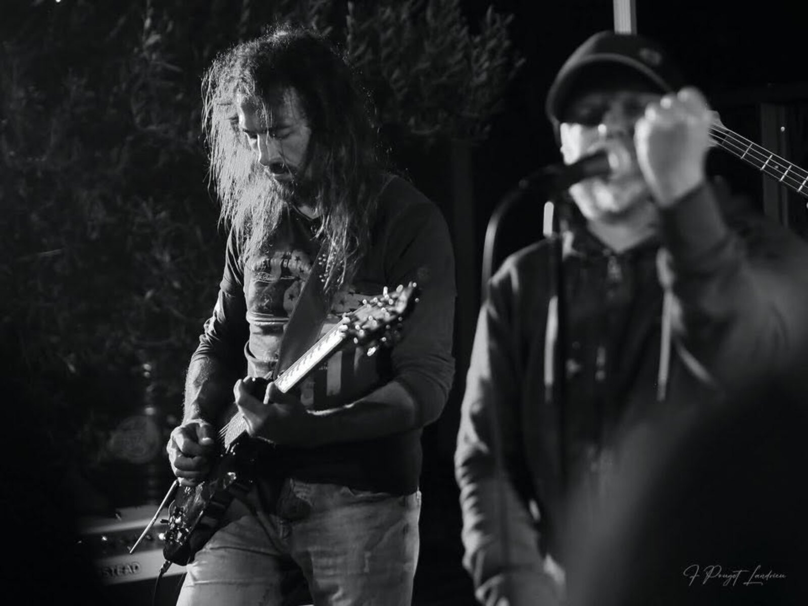 Photo en noir et blanc de Rockface sur scène : un guitariste aux cheveux longs penché sur son instrument, et au second plan le chanteur casquette vissée sur la tête, le poing serré sur le micro