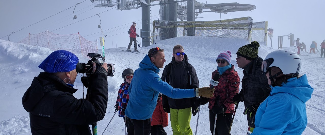 2018, Serge, bénévole, interviewe un groupe de skieurs au sommet des Sybelles, caméraman au premier plan, télésiège en arrière-plan dans le brouillard.