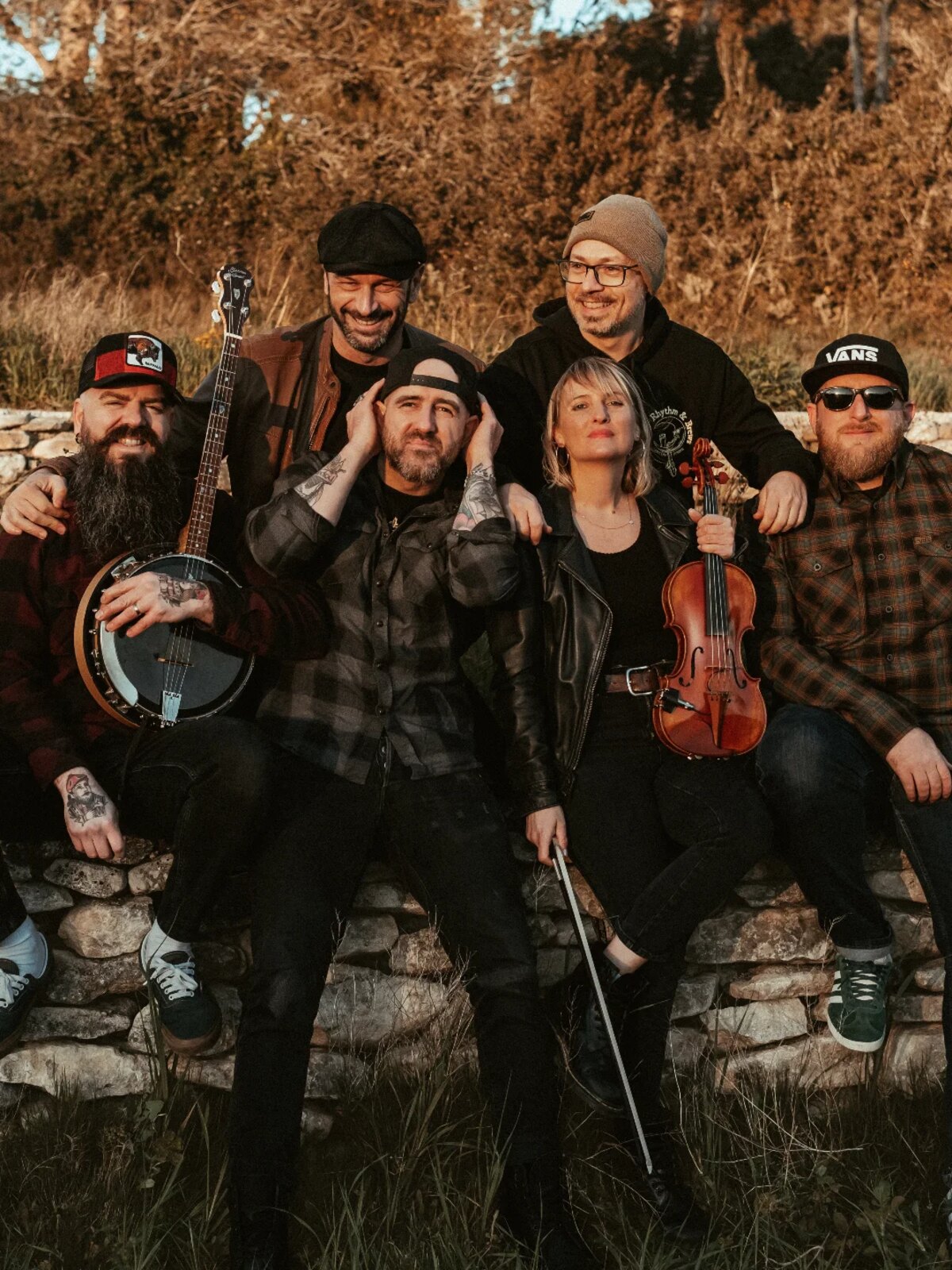 Portrait des six musiciens du groupe de rock celtique The Mullins, assis et debout sur un muret de pierre dans la lumière dorée de l'automne, avec un banjo et un violon