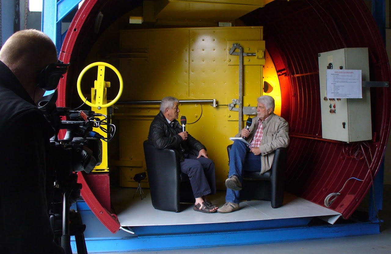 2013, Jean-Claude en interview dans une installation industrielle, structure cylindrique rouge et jaune, micros à la main.