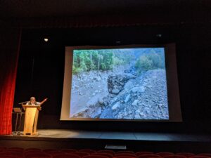 Alain Marnezy au pupitre, le bras tendu vers la photo projetée d'un lit de torrent creusé dans les pierres et les blocs, entre des versants boisés