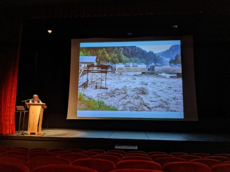Alain Marnezy au pupitre sur une scène de théâtre, devant la projection d'une rivière en crue aux eaux boueuses au pied d'un barrage