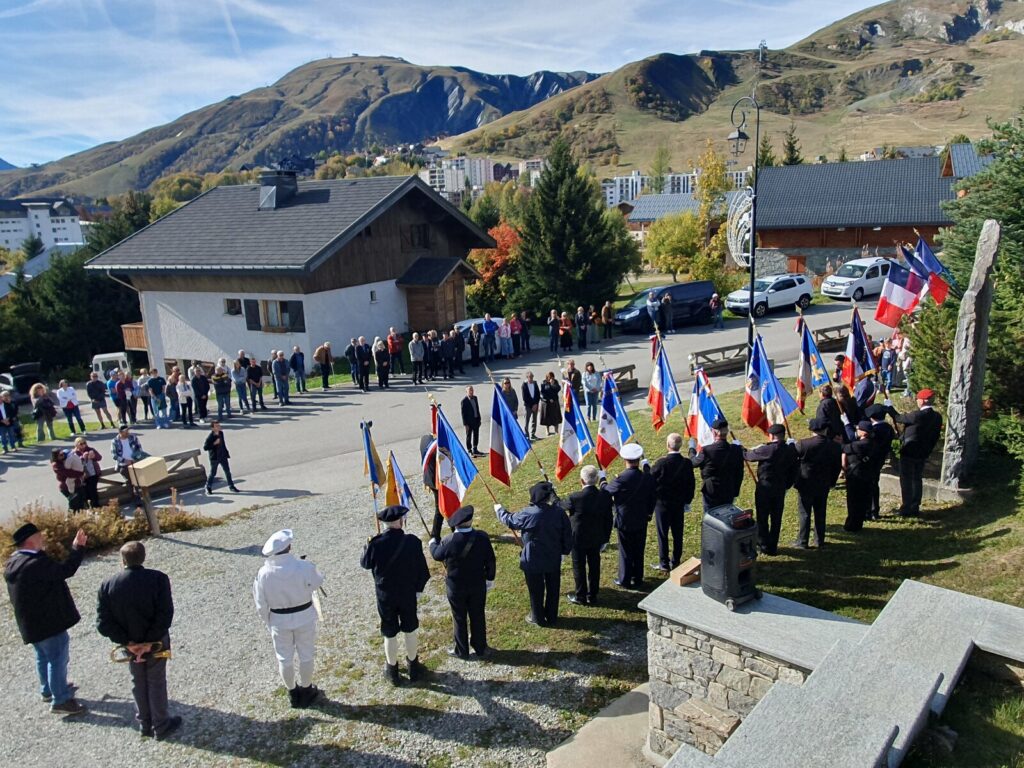 Vue en plongée sur la cérémonie : la ligne des porte-drapeaux de dos face à la stèle, le public massé le long de la rue, les immeubles de la station et les montagnes de La Toussuire en arrière-plan