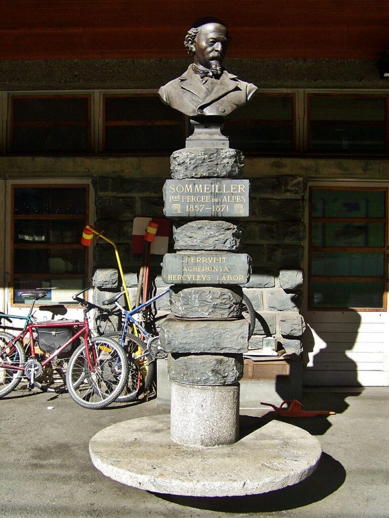 Buste en bronze de Germain Sommeiller sur une colonne de blocs de pierre dans la gare de Modane, avec la plaque « Sommeiller, 1ère percée des Alpes, 1857-1871 » et une devise latine