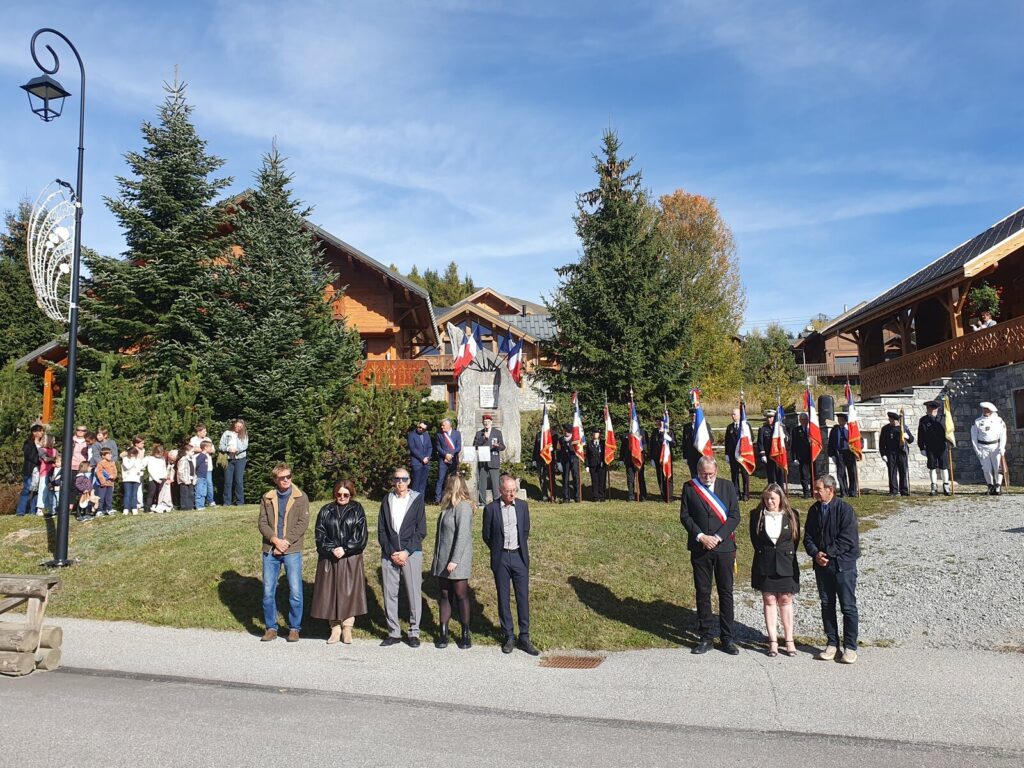 Vue d'ensemble de la cérémonie : élus alignés au premier plan sur le trottoir, porte-drapeaux devant la stèle, enfants des écoles rassemblés sous un sapin à gauche, chalets de La Toussuire et ciel bleu