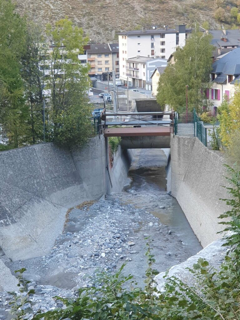 Chenal en béton du torrent du Charmaix à Fourneaux, enjambé par une passerelle métallique, avec des immeubles en arrière-plan