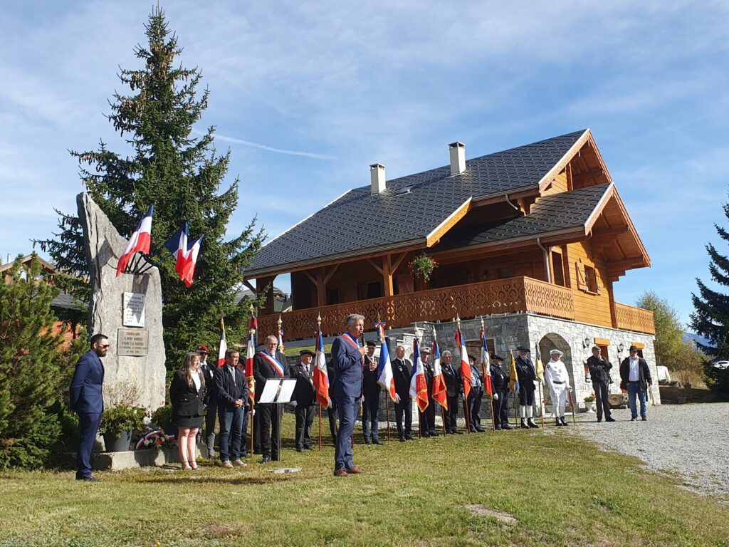 Christophe Truchet, maire de Fontcouverte-La Toussuire, en écharpe tricolore, parle au micro devant la stèle des parachutistes, entouré d'élus et de la ligne des porte-drapeaux, devant un grand chalet de bois
