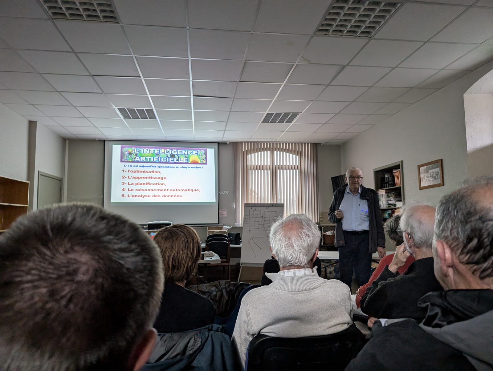 Marc Hivert debout à côté de l'écran qui liste les cinq branches de l'IA : optimisation, apprentissage, planification, raisonnement automatique, analyse des données