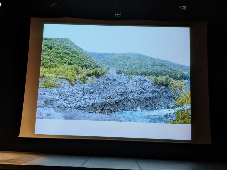 Diapositive projetée : un épais dépôt de pierres et de boue laissé par une lave torrentielle au bord d'une rivière, entre des versants boisés