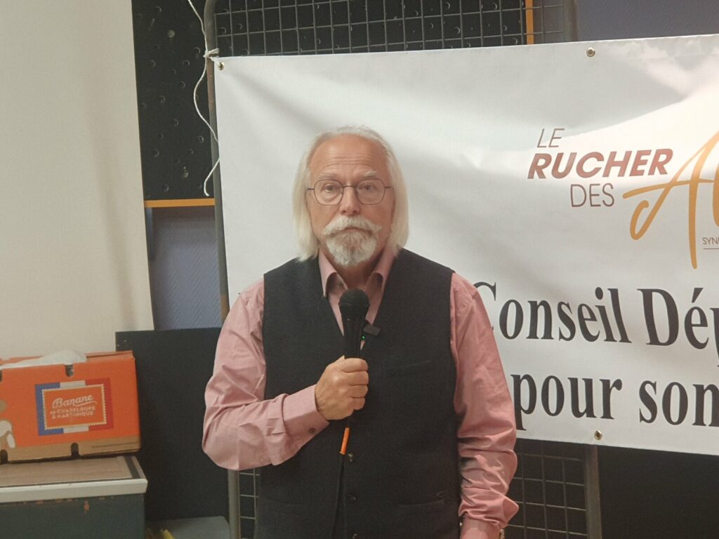 Portrait de Joseph Hemmerlé, cheveux blancs et bouc, chemise rose et gilet sombre, un micro à la main devant la banderole du Rucher des Allobroges