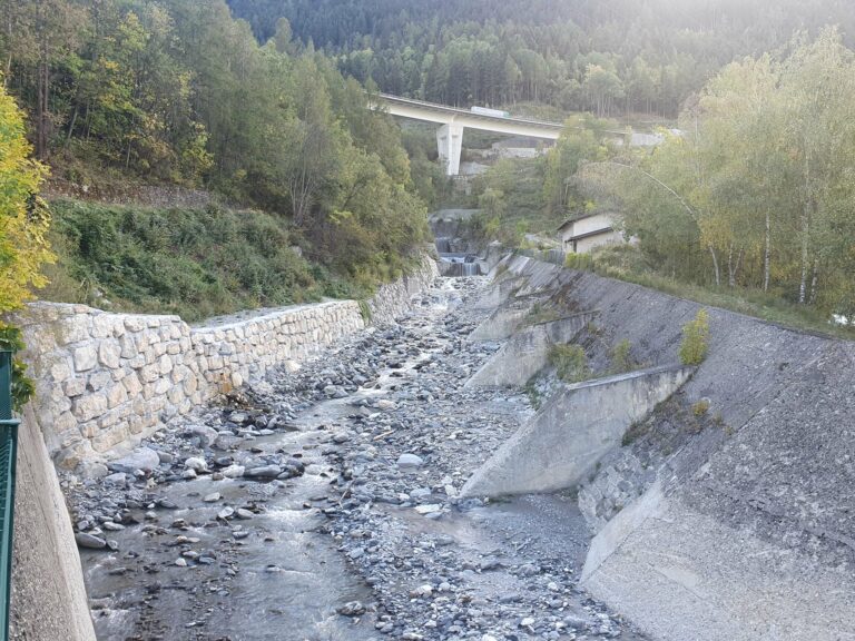 Lit de galets du torrent du Charmaix entre un mur en pierres et des berges bétonnées, un viaduc routier en arrière-plan