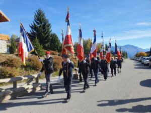 Défilé des porte-drapeaux en costume sombre et gants blancs, drapeaux tricolores levés, le long de la rue des Chasseurs Alpins à La Toussuire, montagnes en arrière-plan