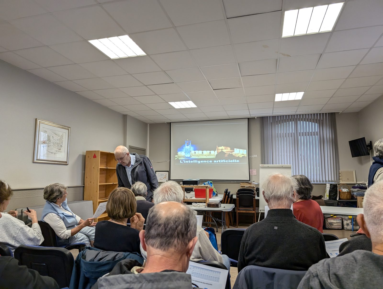 Salle de spectateurs assis face à Marc Hivert, debout près de l'écran de projection titré « L'intelligence artificielle »