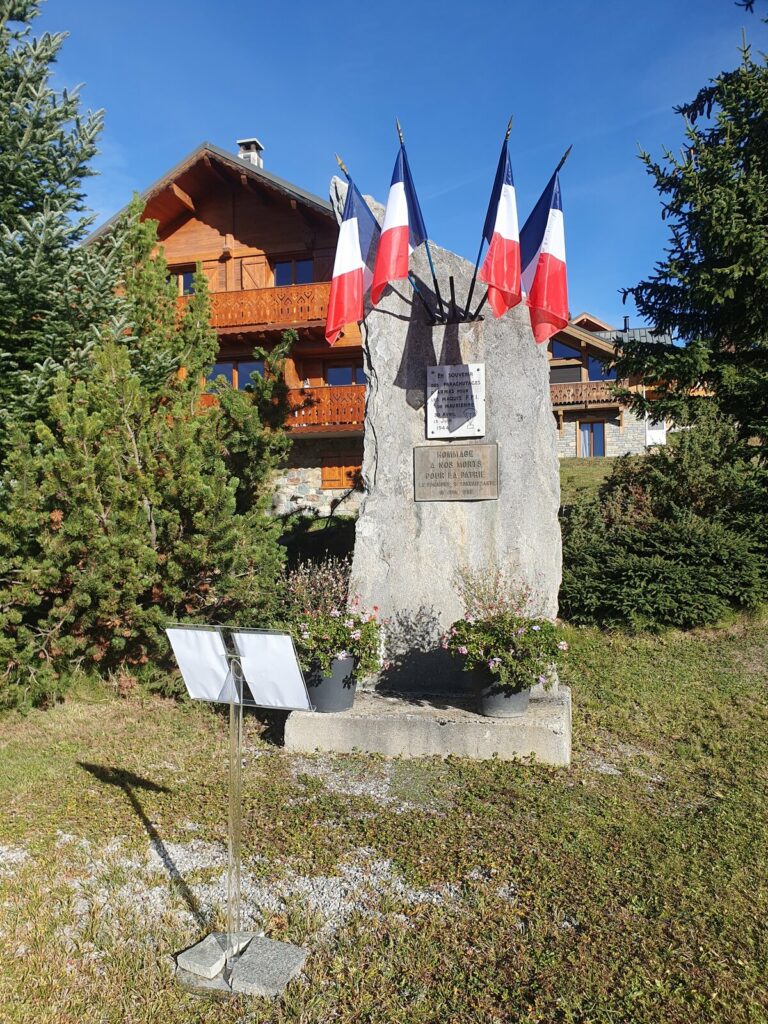 La stèle des parachutistes de La Toussuire : un rocher dressé portant une plaque blanche et une plaque de bronze, cinq drapeaux tricolores au sommet, deux jardinières fleuries au pied et un pupitre devant, un chalet en arrière-plan