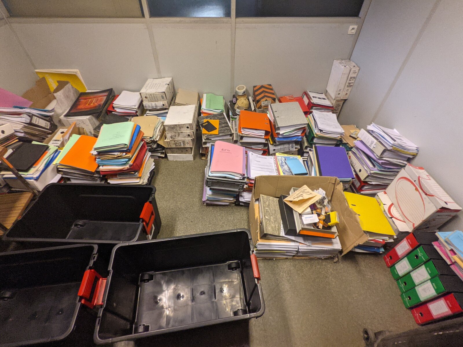 Piles de dossiers et de classeurs de couleurs triés au sol, avec des bacs de transport, lors du déplacement des archives en 2021